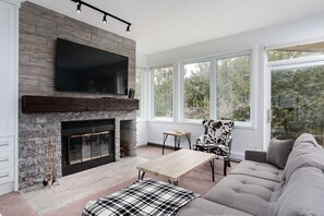 A modern living room with a fireplace, a flat-screen TV mounted above it, a large window offering a view of trees, a sofa, and a coffee table.
