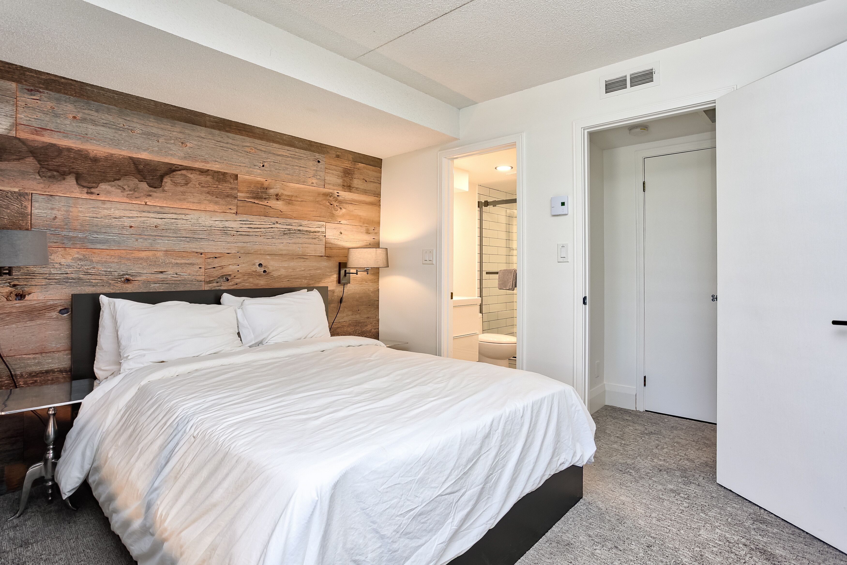 A bedroom with a wooden headboard, a white bedspread, a bedside table with a lamp, and a door leading to a bathroom.