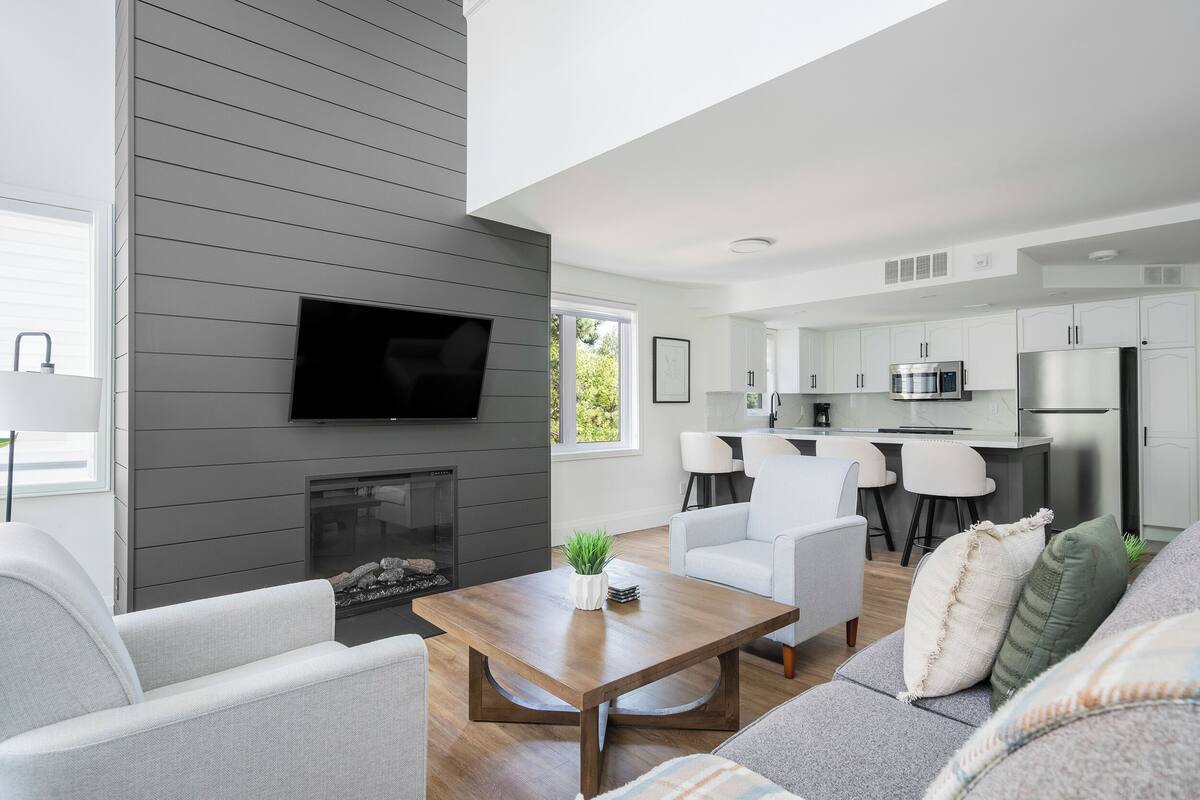 A modern living room with a fireplace, a flat-screen TV mounted on the wall, and a dining area with a kitchen in the background.