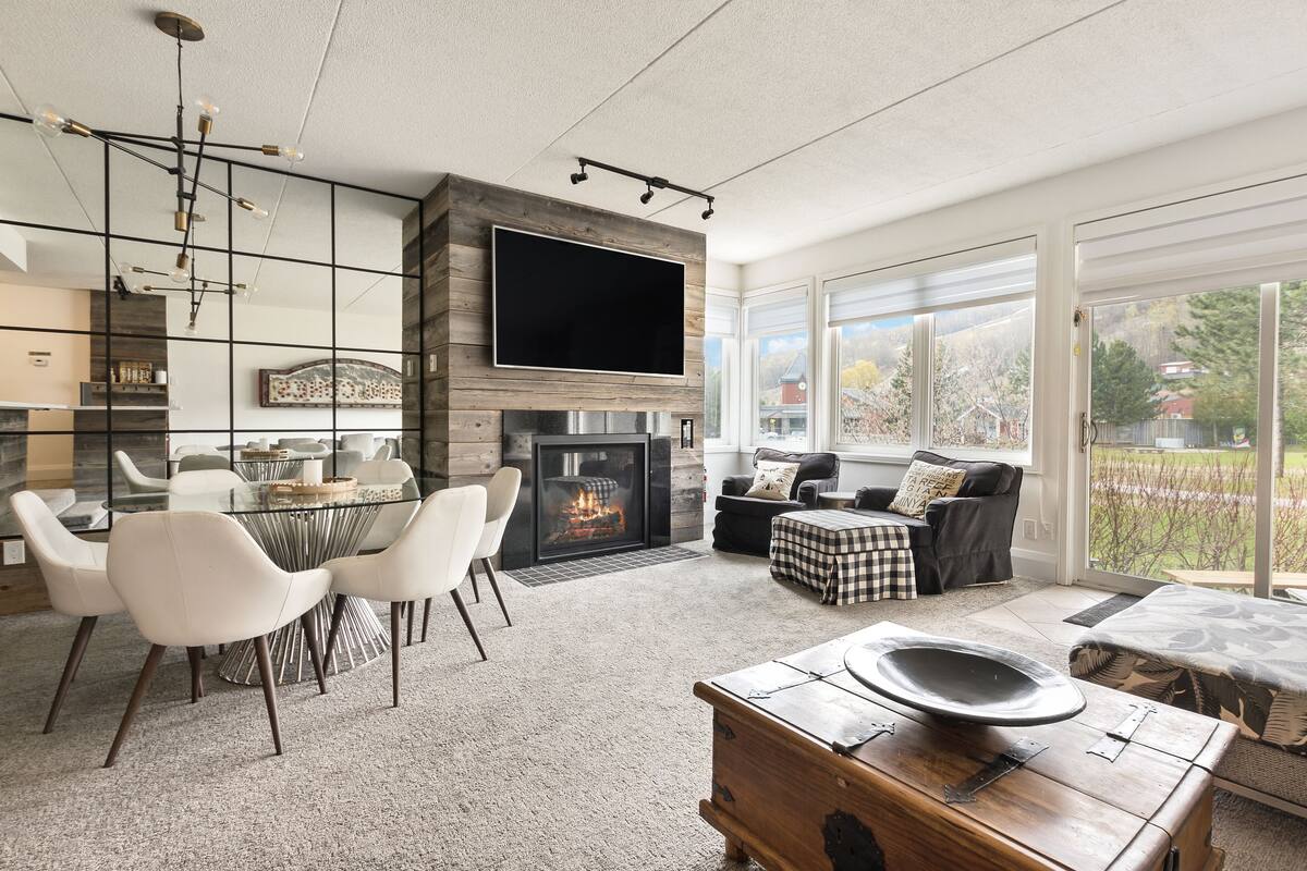 A modern living room with a fireplace, a dining area with chairs, and a large window offering a view of the outdoors.