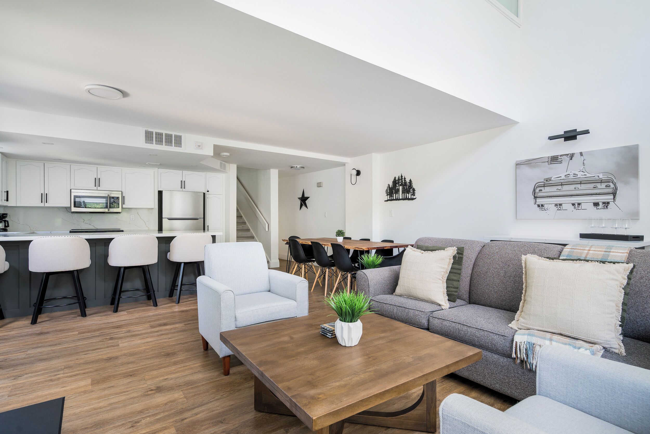 A modern living room with a wooden coffee table, a grey sofa, and a white armchair. There is a kitchen area with white cabinets and a microwave.