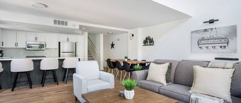 A modern living room with a wooden coffee table, a grey sofa, and a white armchair. There is a kitchen area with white cabinets and a microwave.