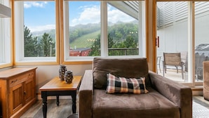 A cozy living room with a brown leather sofa, a plaid pillow, and a wooden table with decorative items. Large windows offer a view of a green landscape and a building in the distance. - Cachet Crossing Chalets (The Blue Mountains)