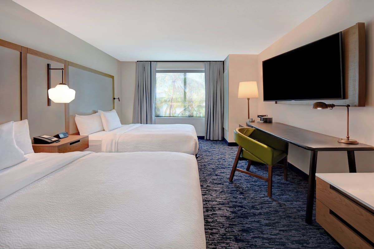A hotel room with two beds, a desk, a TV, and a window with curtains.