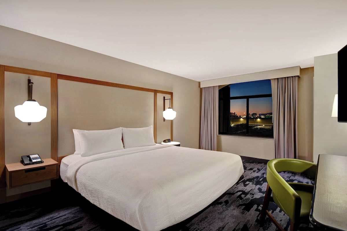 A hotel room with a large bed, bedside tables with lamps, a chair, and a view of the city at dusk.