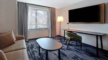 Quarto de hotel com sofá, mesinha, escrivaninha com cadeira, televisão fixada na parede e janela com cortinas.