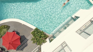 An aerial view of a swimming pool with a person swimming, surrounded by a white building, a red umbrella, and some greenery. - Novotel Suites Manila at Acqua (Mandaluyong)
