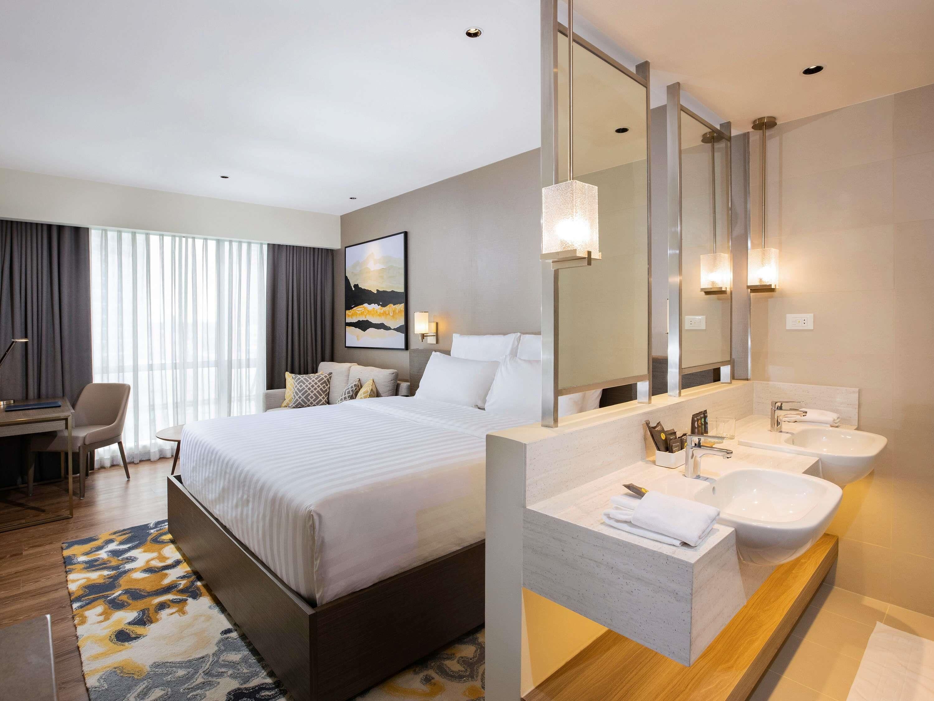 Habitación de hotel con una cama grande, un escritorio, una silla, un baño con lavamanos y espejo, y un ventanal con cortinas.