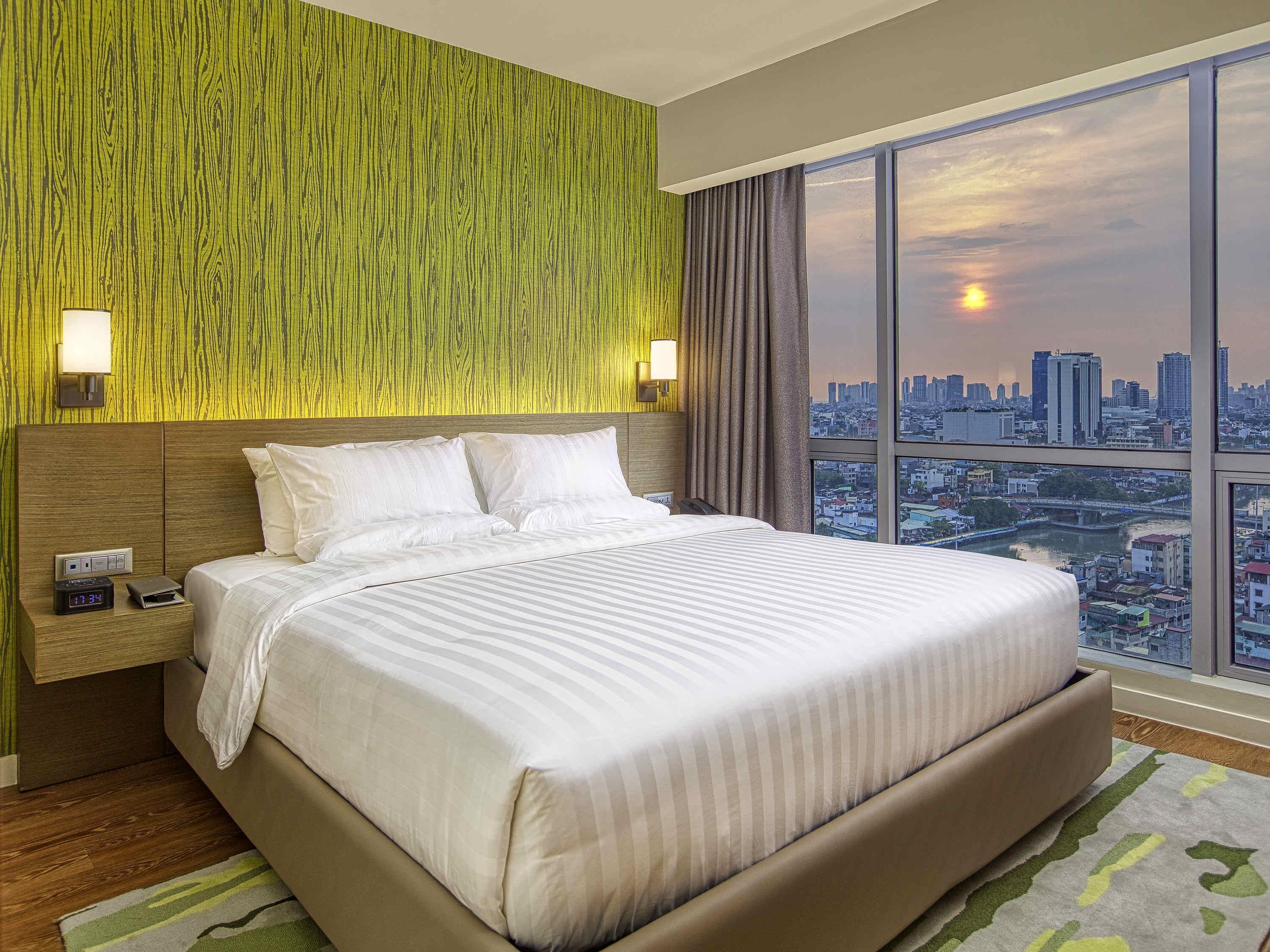 Habitación de hotel con una cama grande, vista a la ciudad, pared de madera y una mesita de noche con reloj.