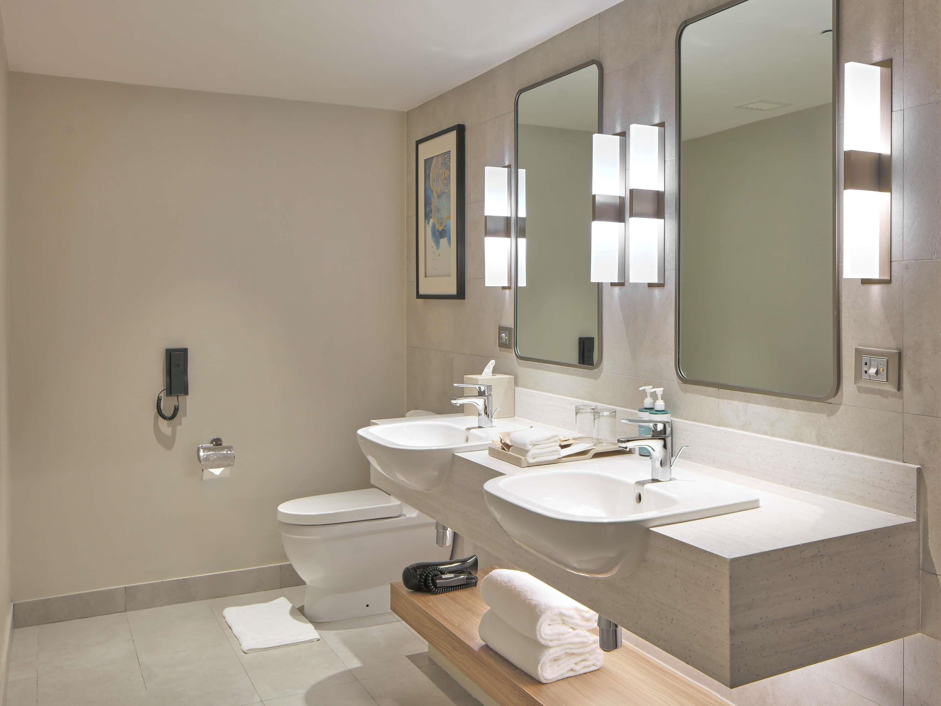 A modern bathroom with a toilet, two sinks, a large mirror, and a towel rack.