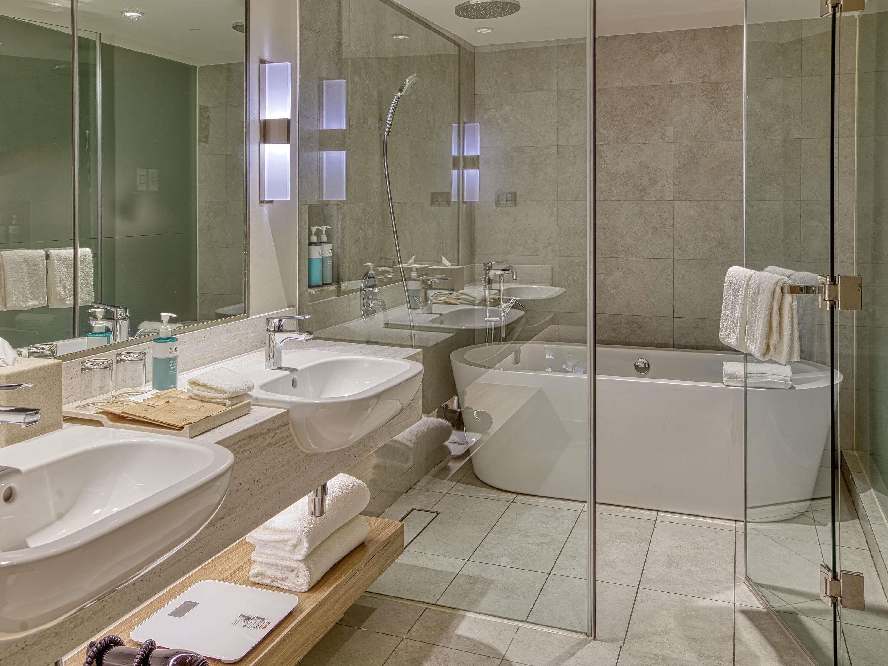 Une salle de bain moderne dotée d’une douche à parois de verre, de deux lavabos, d’une baignoire et d’un meuble-lavabo avec un porte-serviettes.