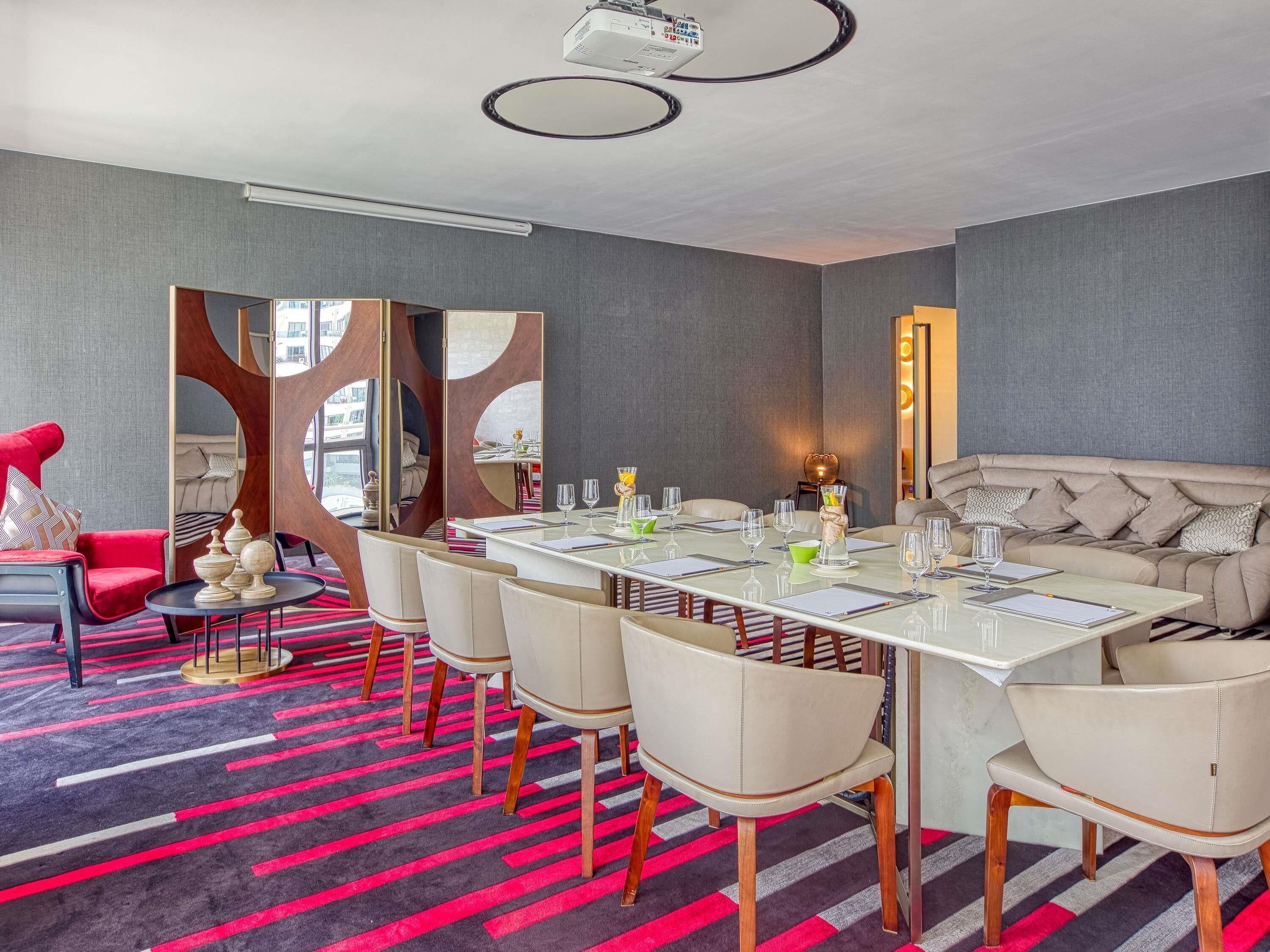 Quarto de hotel moderno com área de refeições, uma poltrona vermelha, um sofá e um espelho grande.
