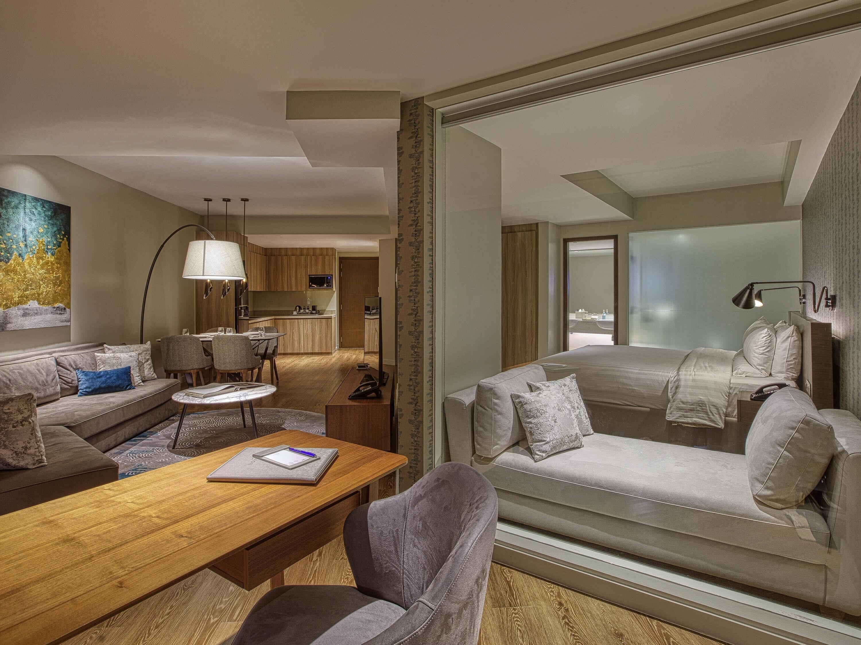 Una habitación de hotel moderna con sofá, una mesa de comedor, una lámpara y vistas a la ciudad a través de un ventanal grande.