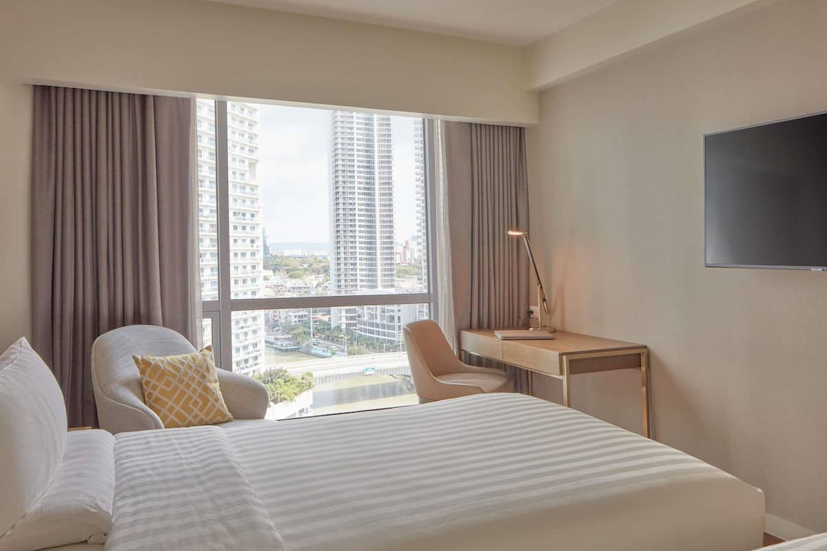 Une chambre d’hôtel avec un lit, deux chaises, un bureau, une télévision et une grande fenêtre avec des rideaux.
