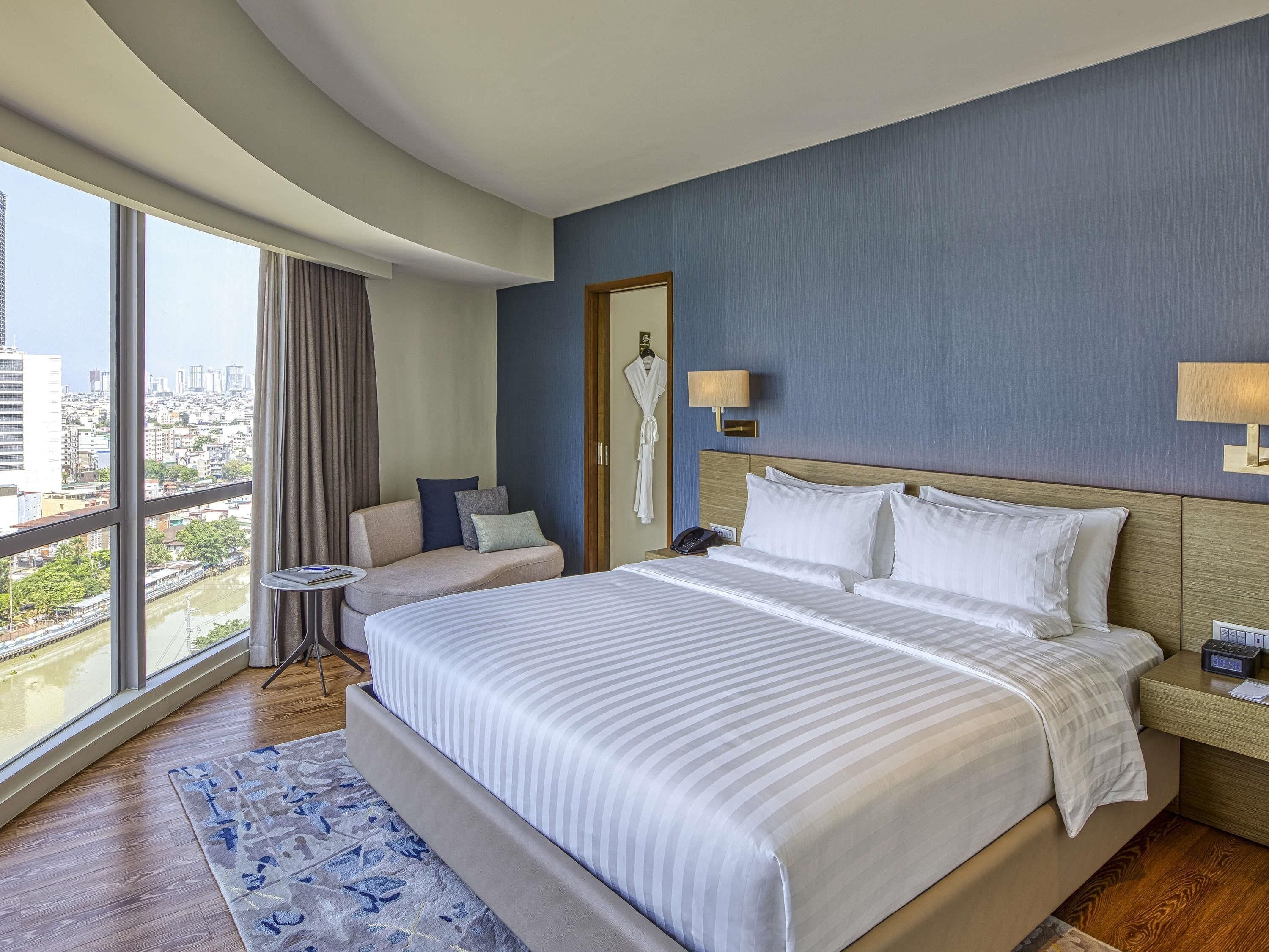 Een hotelkamer met een groot bed, een bank, een kleine tafel en uitzicht op de stad door grote ramen.