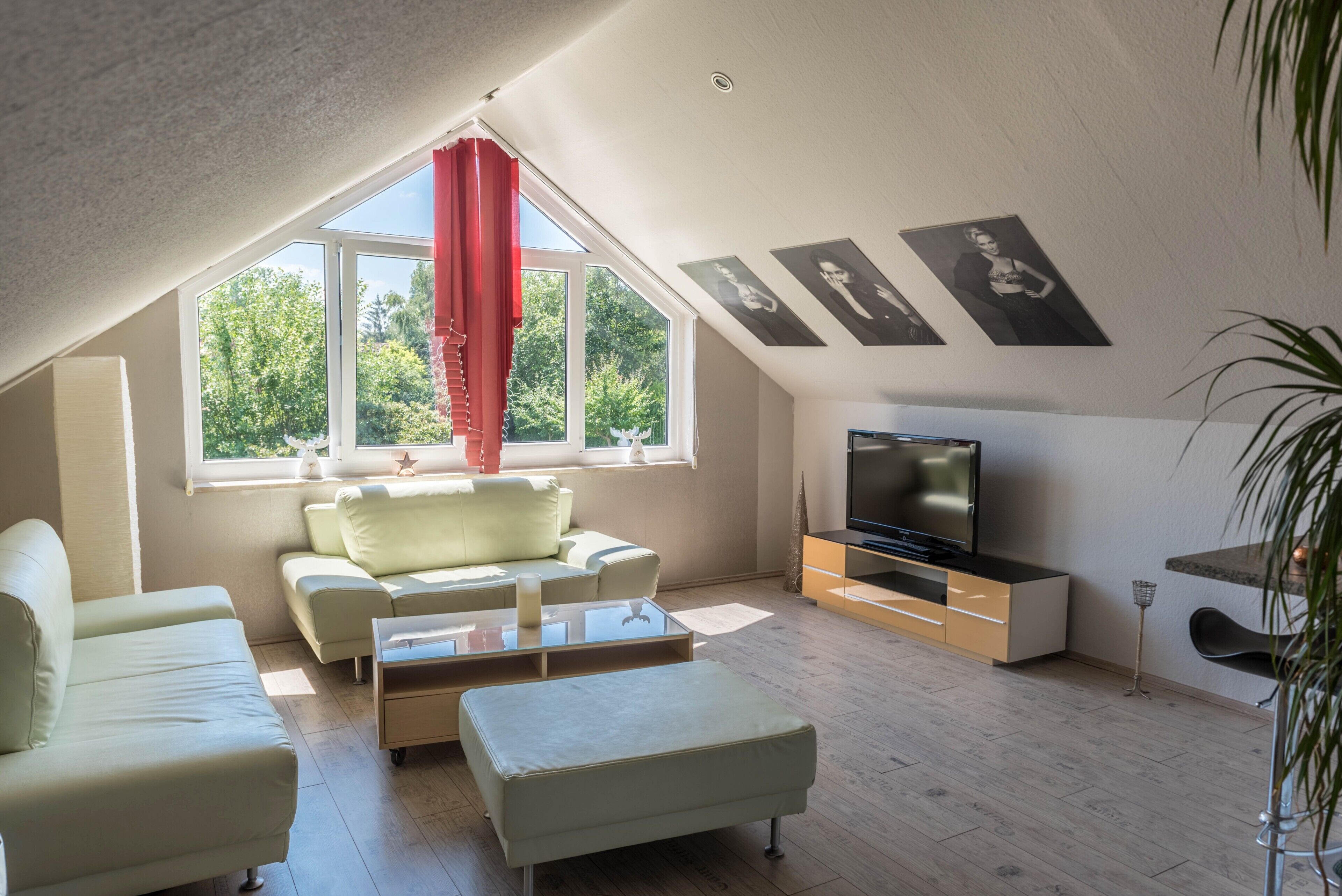 A modern living room with a sloped ceiling, a large window with red curtains, a flat-screen TV, and a sofa.