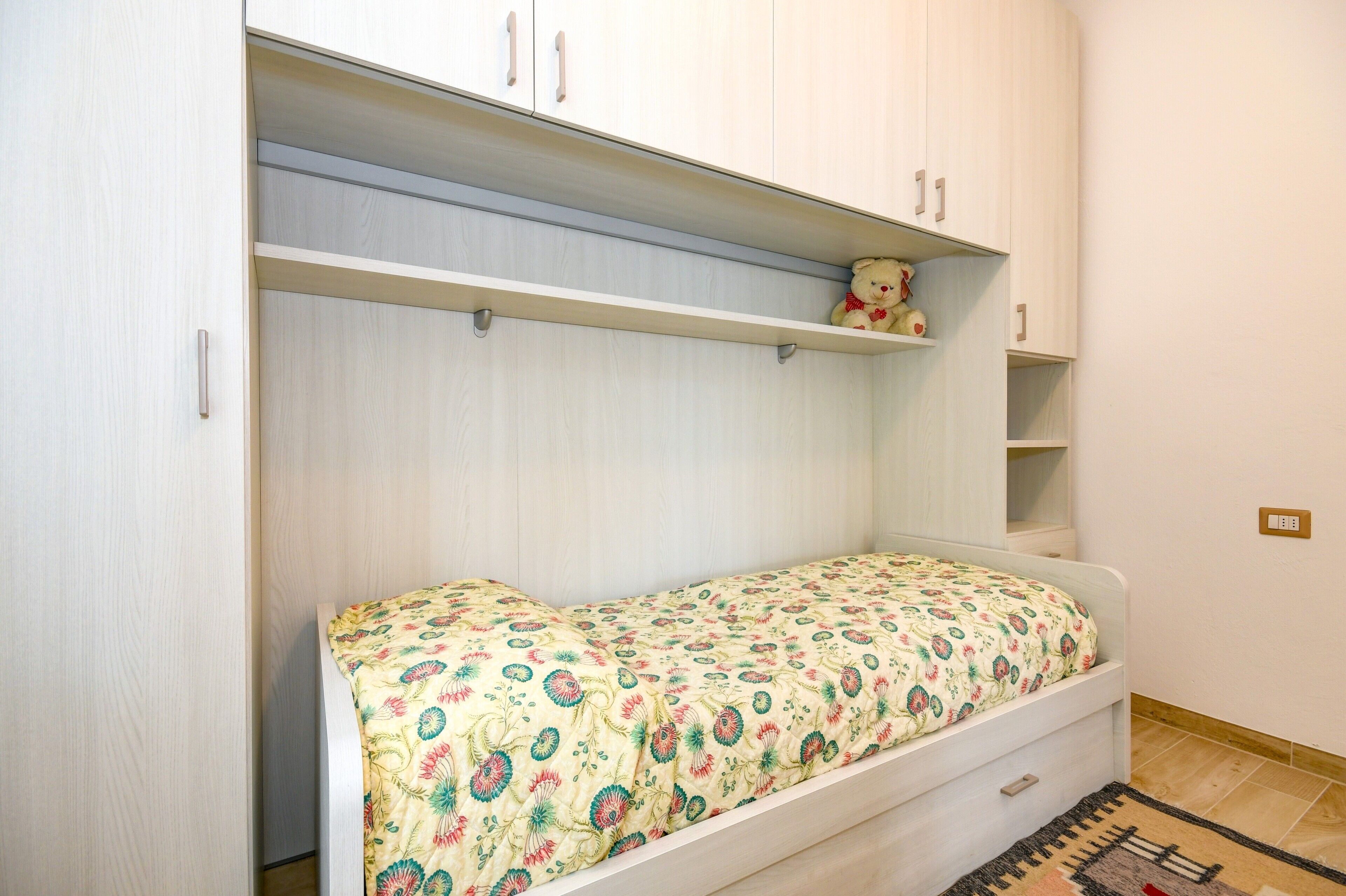 A single bed with a floral bedspread, a small shelf with a teddy bear, and a wardrobe with two doors.