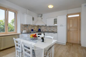 A kitchen with white cabinets, a tiled backsplash, a dining table with chairs, and a window with a view of trees.