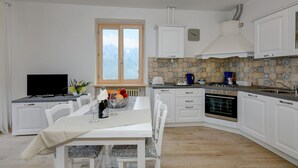 A modern kitchen with white cabinetry, a dining table with chairs, a television, and a window with a view of mountains. - Casa Doss Apartment with Lake View, Private Terrace, and Wi-Fi (TREMOSINE)