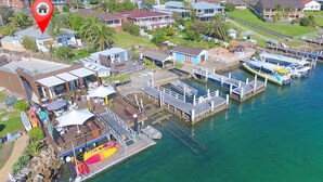 Vue aérienne d’une marina avec des bateaux amarrés, un restaurant doté d’une terrasse, et des immeubles résidentiels en arrière-plan.