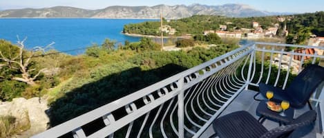 A balcony with a view of the sea and mountains, a chair, and a table with drinks.