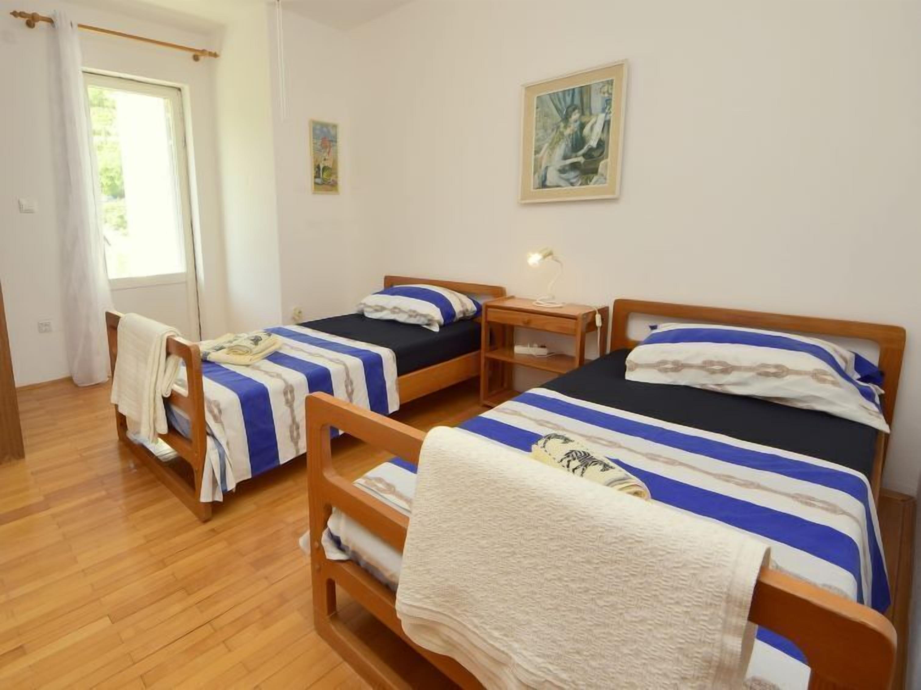 Une chambre avec deux lits en bois, chacun recouvert d’une couverture à rayures bleues et blanches. Une petite table de chevet en bois avec une lampe se trouve entre les deux lits.