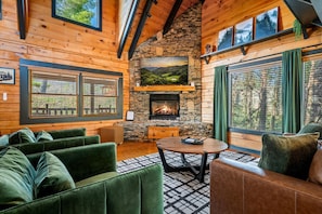 A cozy cabin interior with a stone fireplace, green upholstered seating, and large windows offering scenic views.