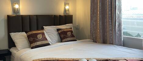 Una cama bien hecha con sábanas blancas y almohadones decorativos, un cabecero con iluminación integrada, y vistas de la ciudad a través de una ventana con cortinas.