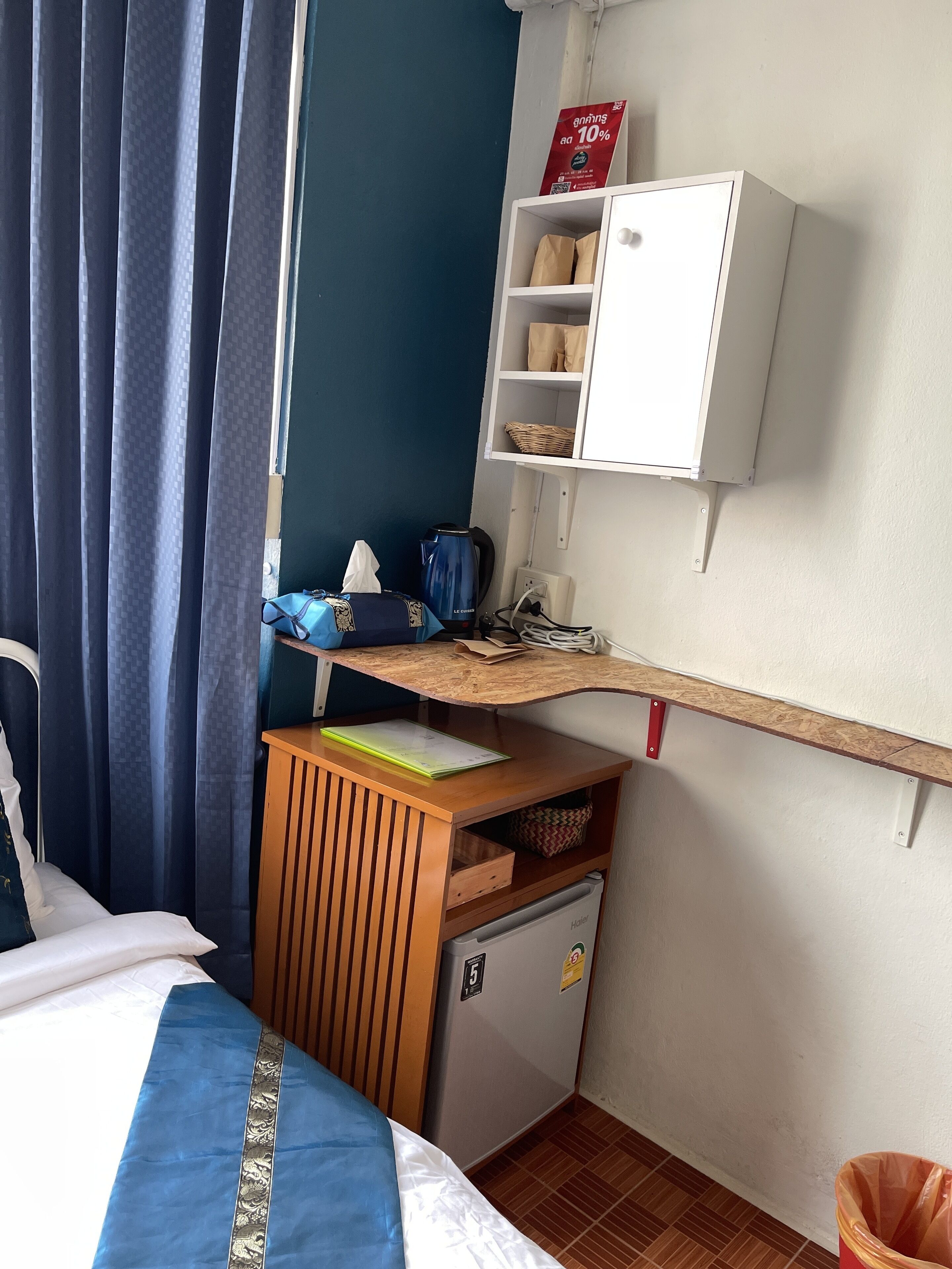 A hotel room with a bed, a small desk with a kettle, a cabinet, and a trash bin.