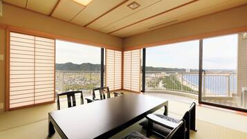 Una habitación con suelo de tatami, una mesa baja y sillas. Amplias ventanas ofrecen vistas a un paisaje urbano y montañas.