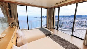 Habitación de hotel con dos camas, un escritorio con teléfono y ventanas amplias que ofrecen vista a la ciudad y al mar.