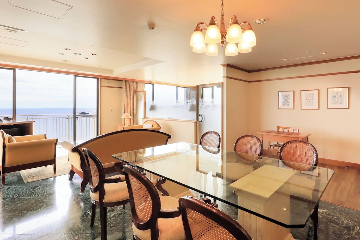 Un vaste espace dédié à la salle à manger, avec une table et des chaises en verre, de grandes fenêtres offrant une vue sur la mer et un lustre.