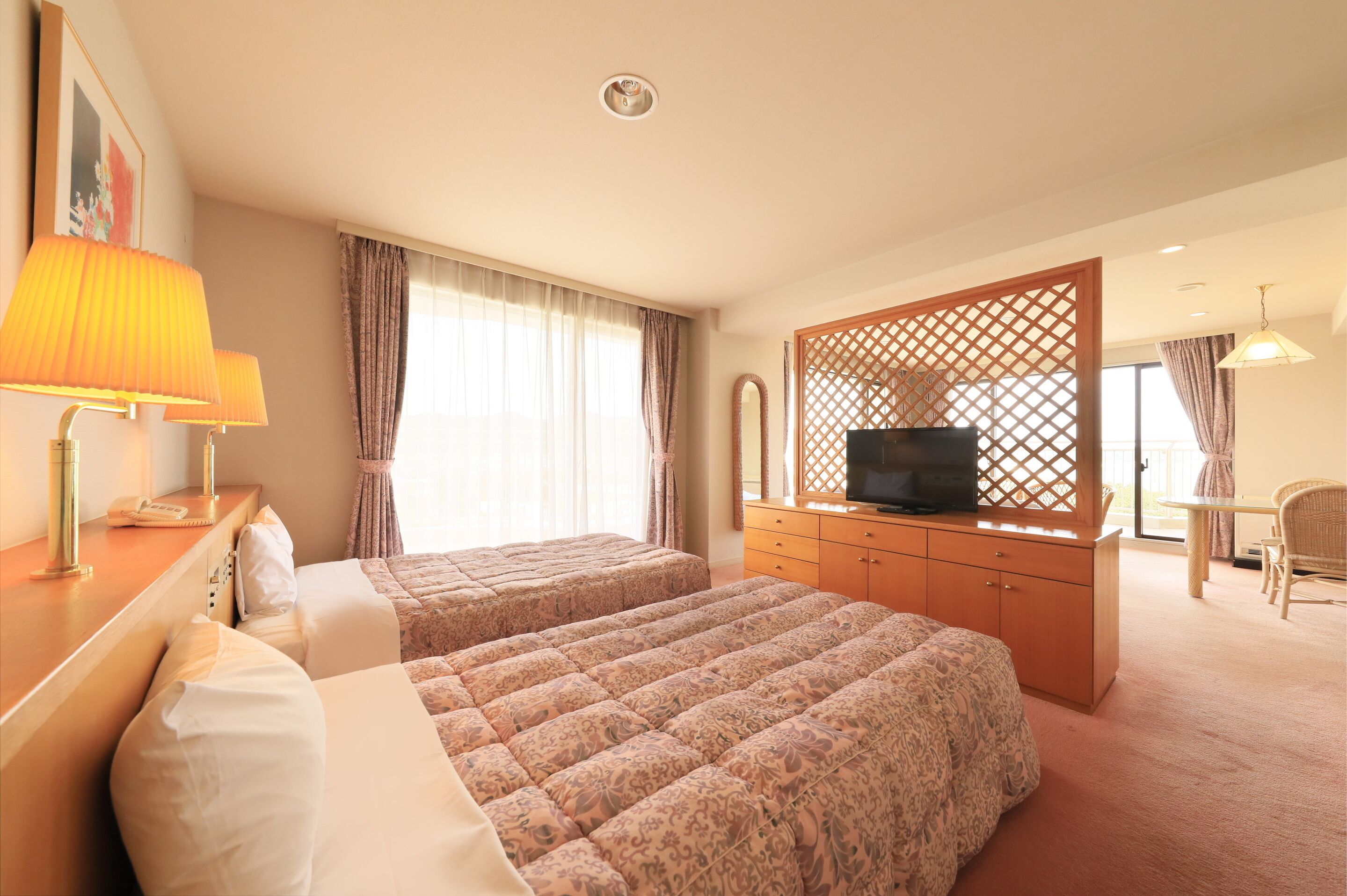 A hotel room with two beds, a TV, a desk with a lamp, and a view of the outside through large windows.