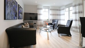 Un salon moderne avec un canapé en cuir, une table basse en verre et un fauteuil en cuir noir. Un téléviseur à écran plat est fixé au mur, et des tableaux abstraits sont accrochés au-dessus.