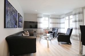 Uma sala de estar moderna com um sofá de couro, uma mesa de centro de vidro e uma poltrona de couro preta. Uma TV de tela plana está fixada na parede, e há quadros abstratos acima.