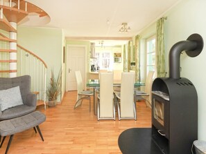 A living room with a wooden spiral staircase, a black wood stove, a dining area with a glass table, and a kitchen in the background.