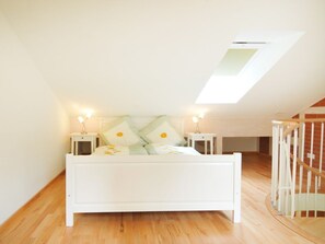 Una cama blanca en una habitación con techo inclinado, suelo de madera y tragaluz.