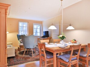 Un comedor con una mesa de madera, sillas y una araña. Se observa un televisor y una ventana al fondo.