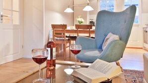Un salon moderne avec un fauteuil bleu, une table en verre sur laquelle se trouvent un livre et deux verres à vin, et un coin repas à l’arrière-plan.