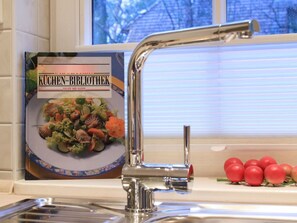 Une cuisine dotée d’un robinet en acier inoxydable, d’un livre de recettes intitulé « KUCHEN-BIBLIOTHEK SALATE DER SAISON » posé sur le comptoir, et d’une fenêtre avec des stores.