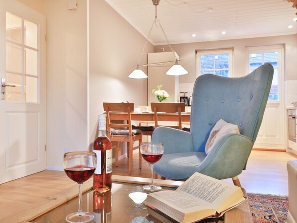 A modern living room with a blue armchair, a glass table with a book and two wine glasses, and a dining area in the background.