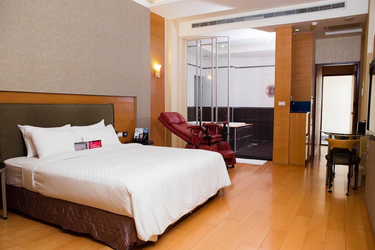 Ein Hotelzimmer mit einem großen Bett, einem roten Liegesessel, einem kleinen Tisch und Blick durch eine Glastür nach draußen.