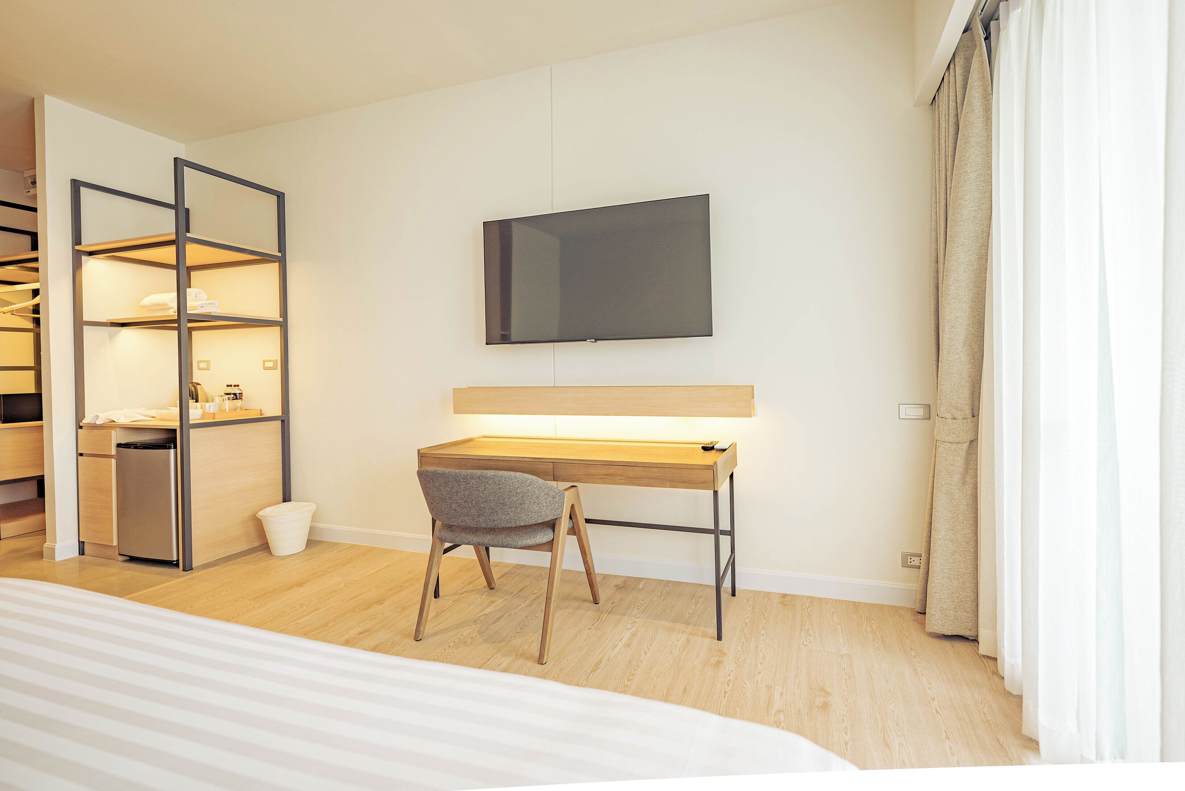 A hotel room with a bed, a desk with a chair, a TV mounted on the wall, a wardrobe, and a small kitchenette.