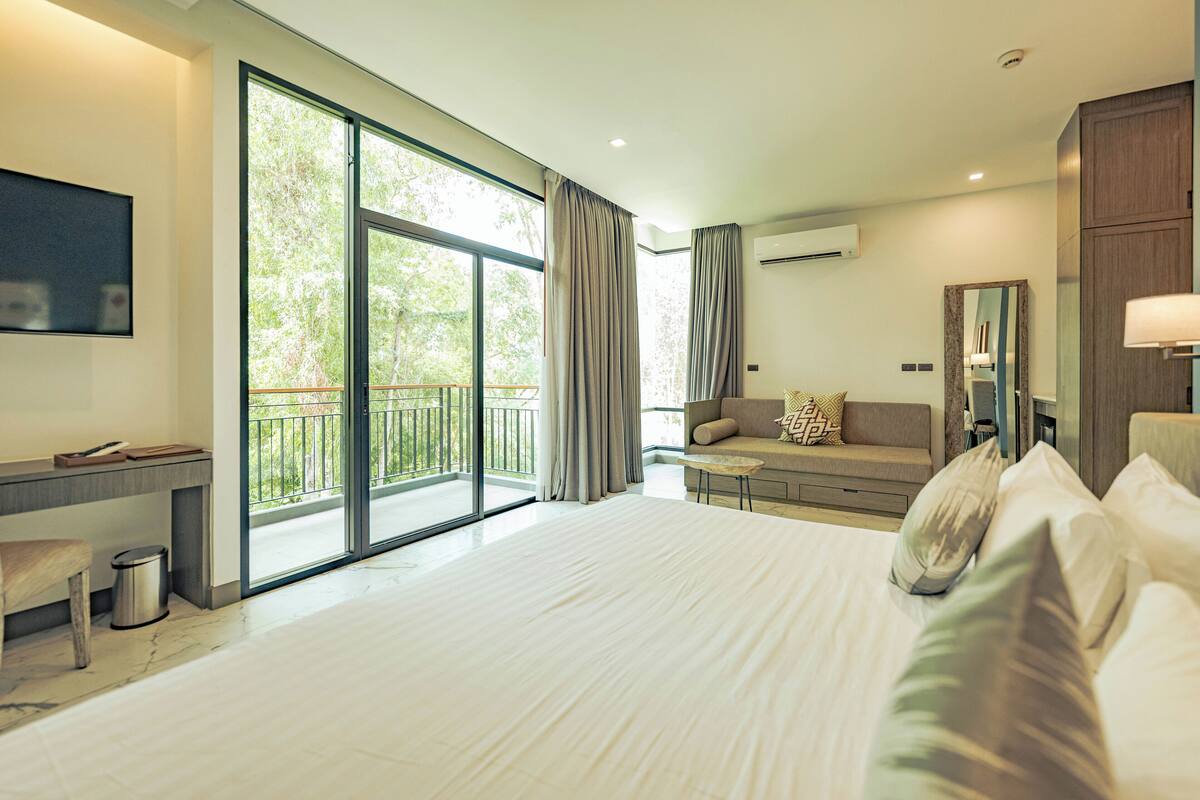 A modern hotel room with a large bed, a sofa, a desk, and a balcony with curtains.