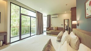 Habitación de hotel con una cama grande, un sofá, una mesita y un balcón con vistas a árboles.