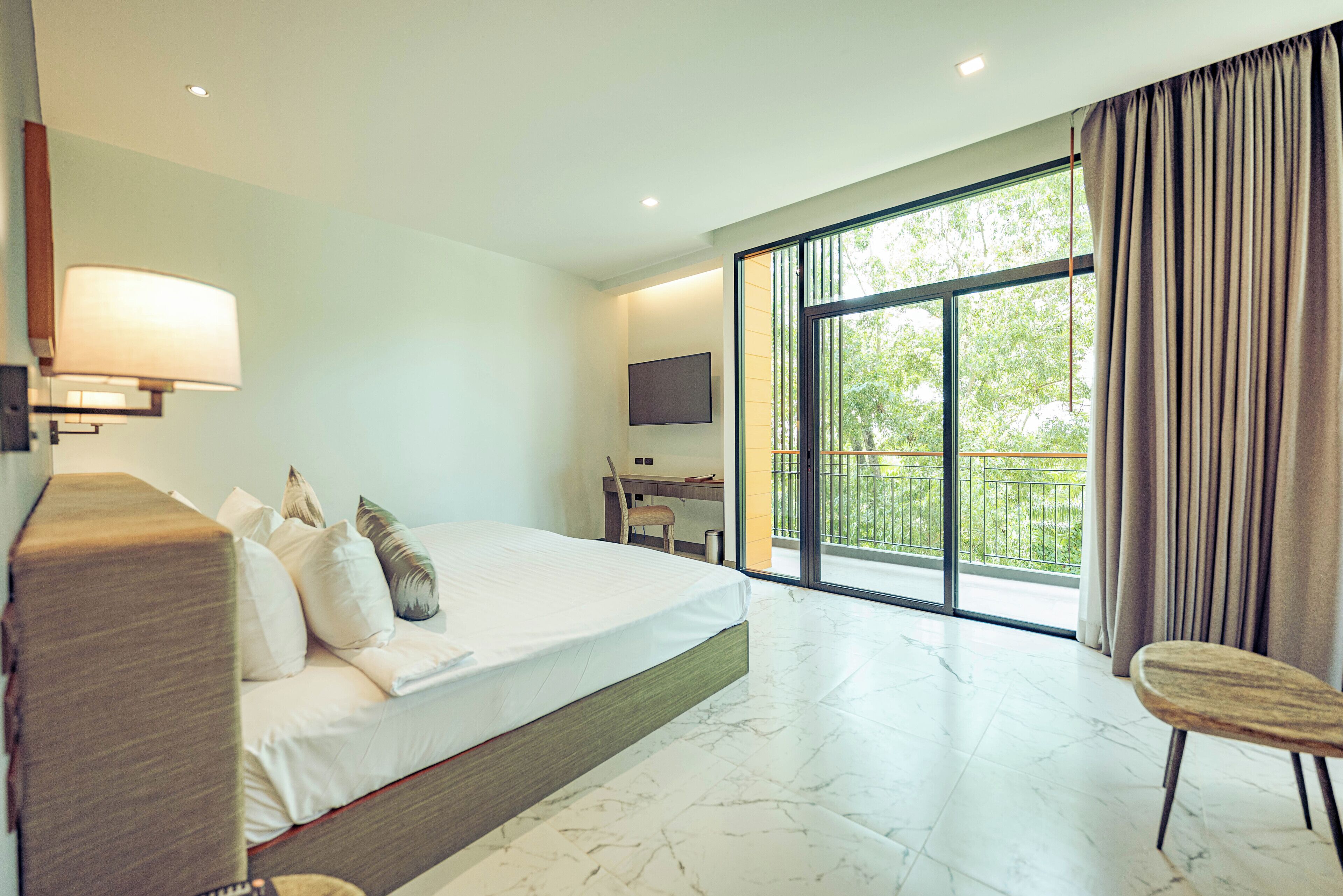 A modern hotel room with a large bed, a desk, a chair, a TV, and a balcony with a view of greenery.
