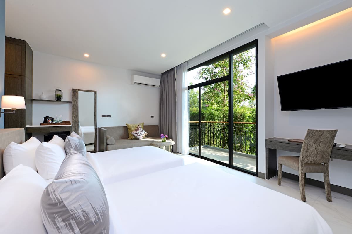 A modern hotel room with a large bed, a desk, a chair, a TV, and a balcony with a view of greenery.