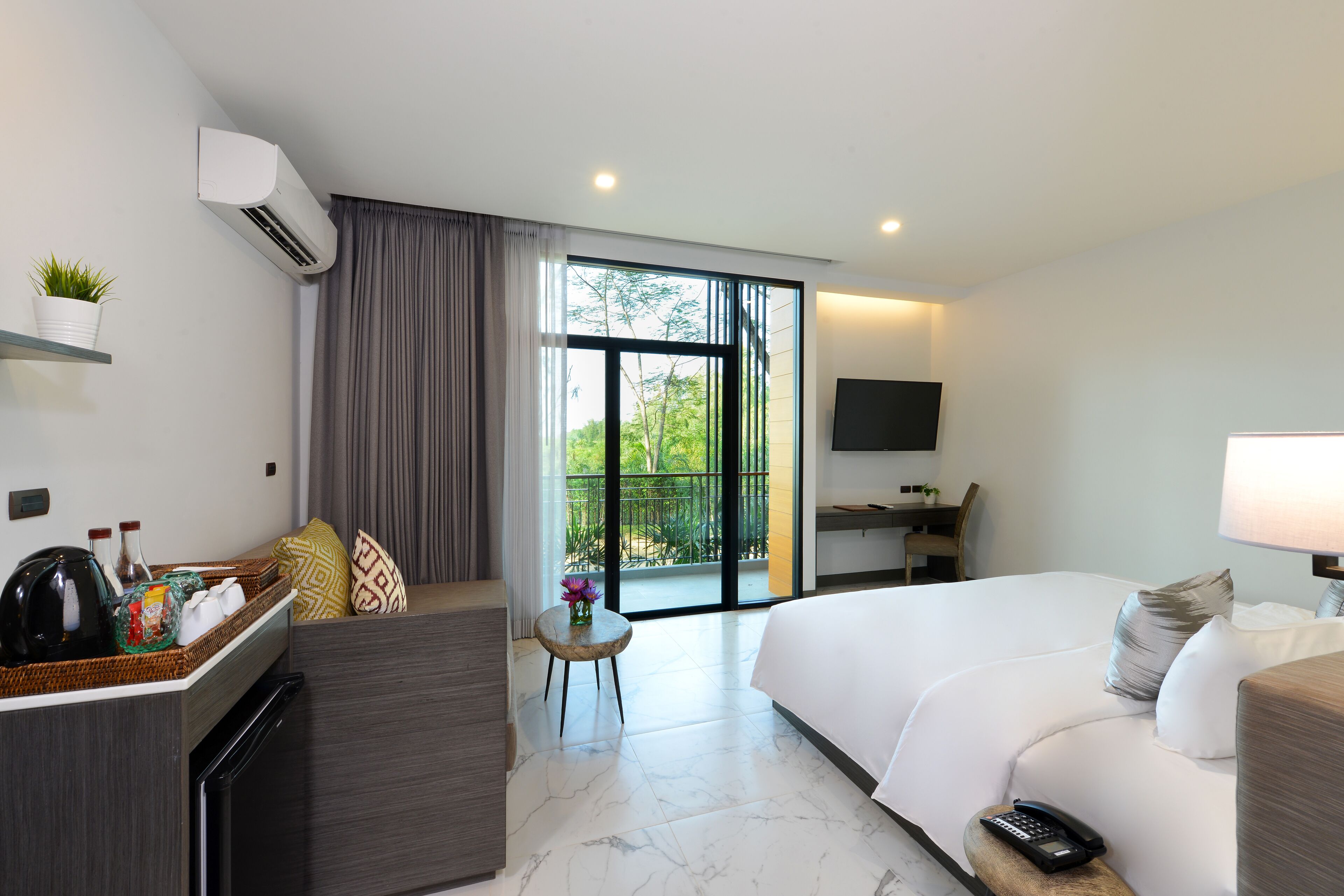 A modern hotel room with a bed, a desk, a small table with a vase, a sofa, and a balcony with a view of greenery.