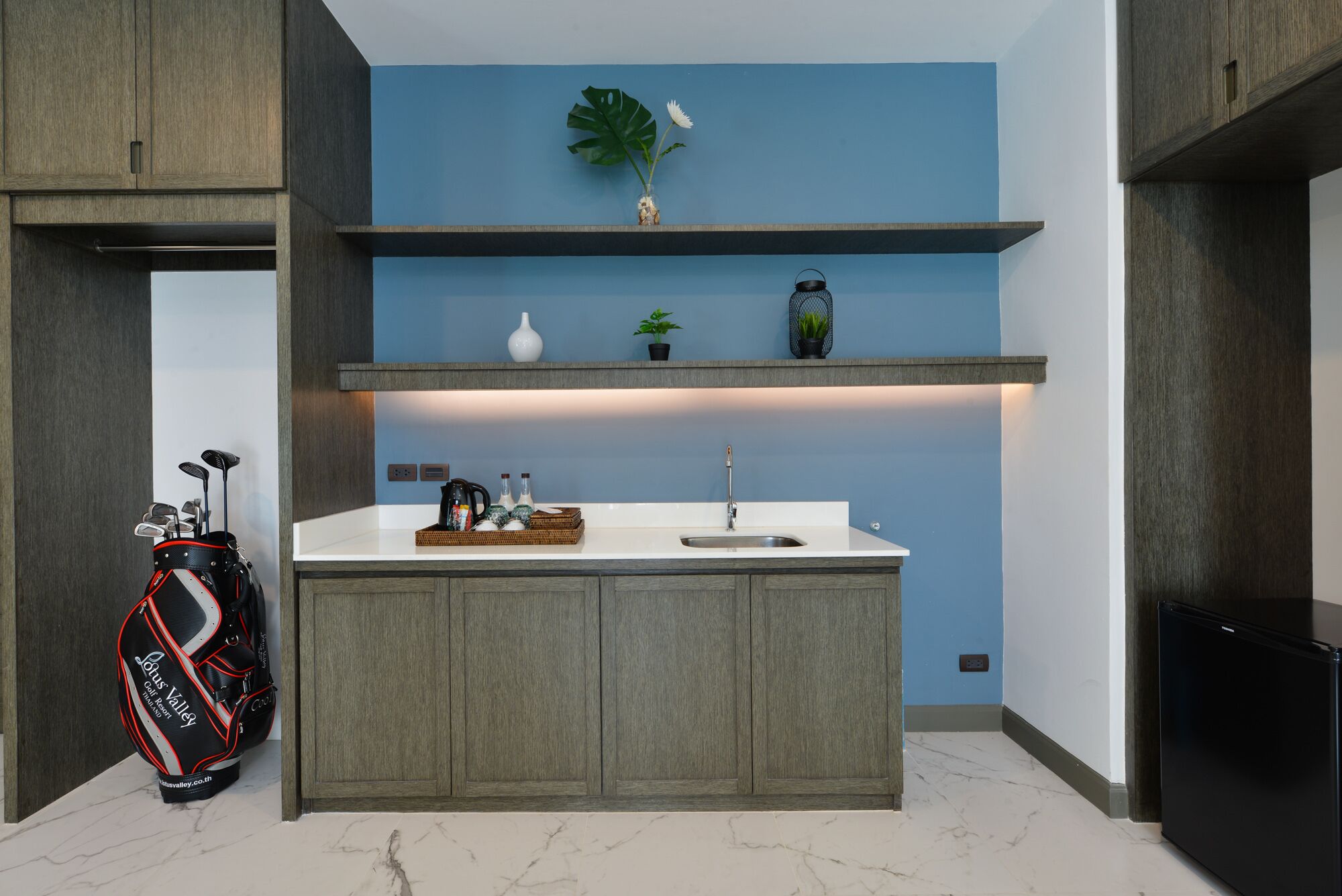 A compact kitchen with a marble countertop, a sink, and a coffee maker. There are two shelves above the countertop, one holding a vase and a plant, and the other with a small potted plant. A golf bag is placed against the wall to the left.