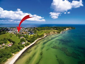 Vista aérea de uma vila costeira, com uma visão nítida da costa, vegetação verde e uma seta vermelha indicando um local específico.